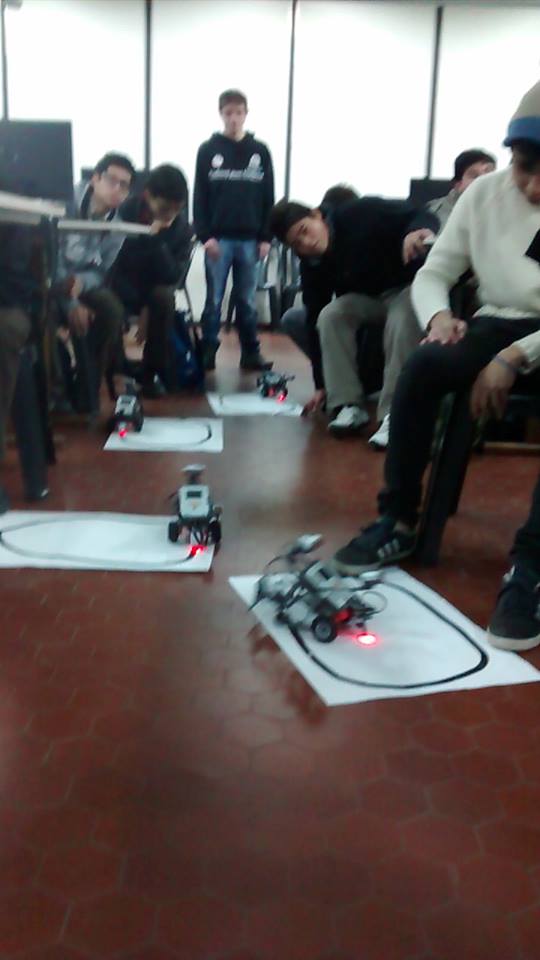 Alumnos de secundaria observando dos robots similares a autos, que andan siguiendo una cinta negra